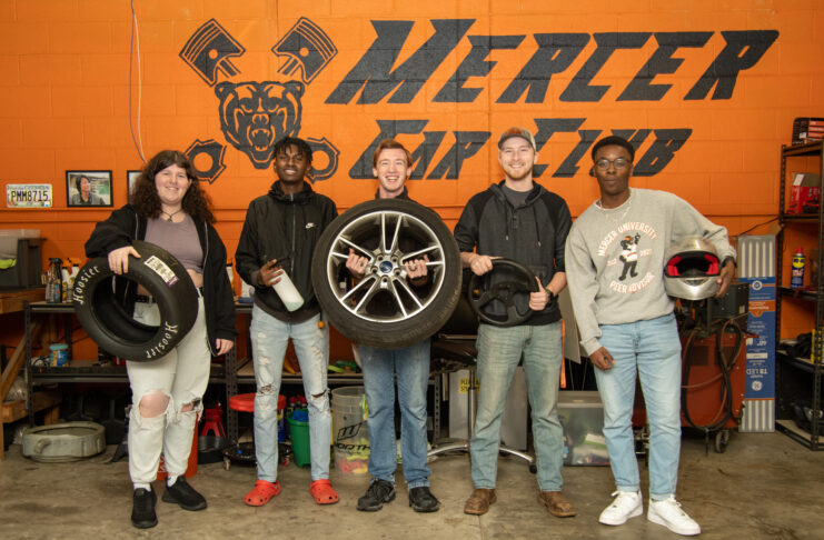 Five students pose for a group photo in the garage, with each holiday a piece of automotive equipment.
