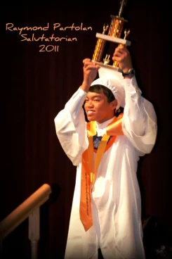 a student in a white cap and gown holds up a trophy