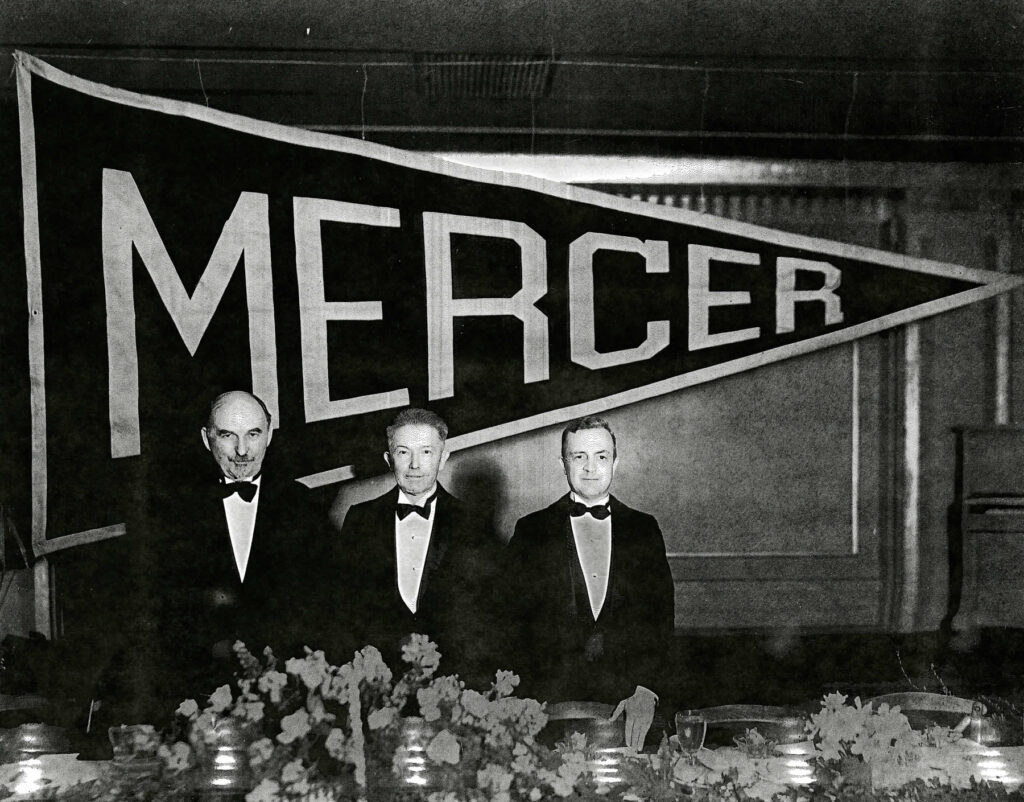 three men stand in front of a large mercer banner