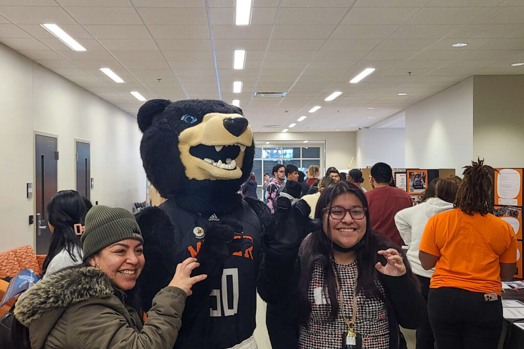 Two people pose for a photo with Bear mascot Toby, while other people look at posters in the background.