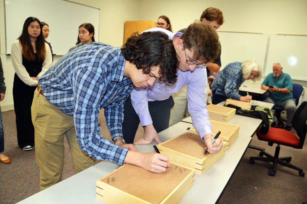 Two students lean over wooden boxes and sign their names.