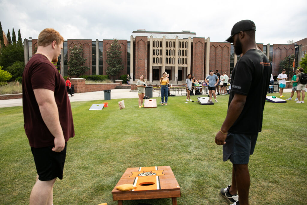 Two rows of cornhole boards are shown on a field with students beside them.