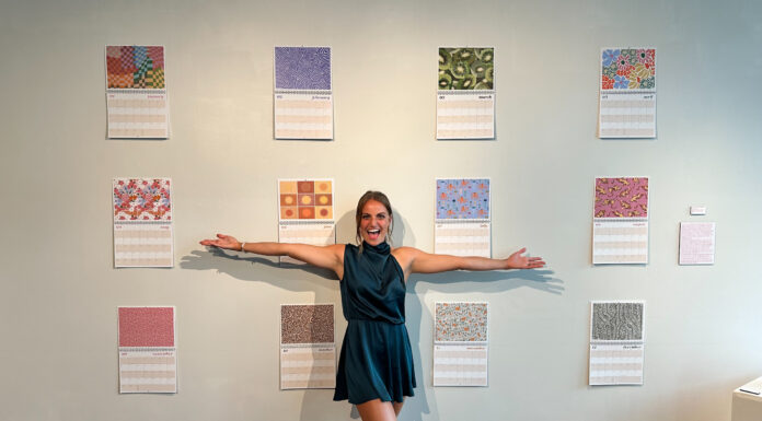 2023 graduate named Graphic Design USA Student to Watch Carly Curran stands with her hands outstretched in front of a wall with 12 calendar pages.