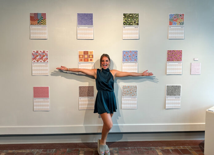 Carly Curran stands with her hands outstretched in front of a wall with 12 calendar pages.