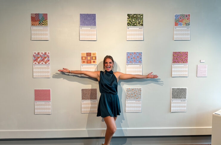 Carly Curran stands with her hands outstretched in front of a wall with 12 calendar pages.