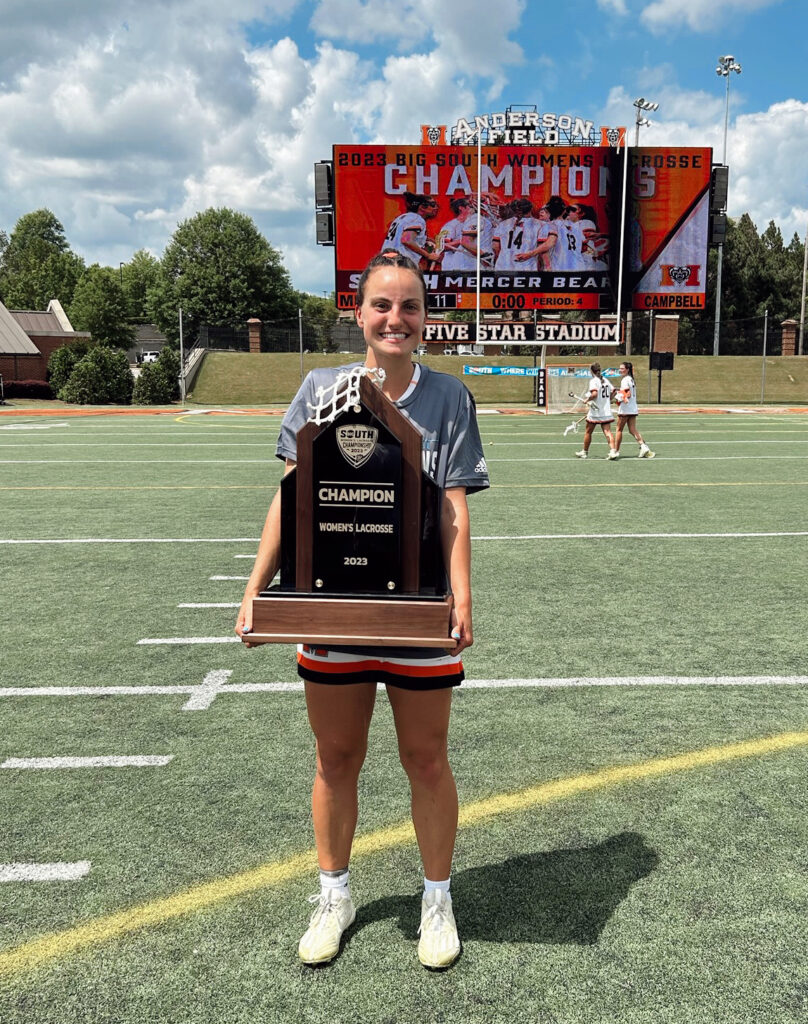 Carly Curran holds a trophy on Anderson Field on Mercer's Macon campus, with a jumbotron in the background that says "2023 Big South Women's Lacrosse Champions."