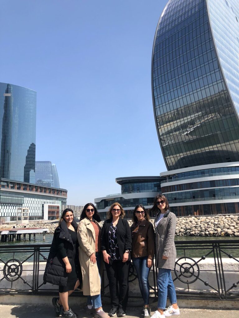 Five women pose in front of a waterfront, with large buildings behind them.