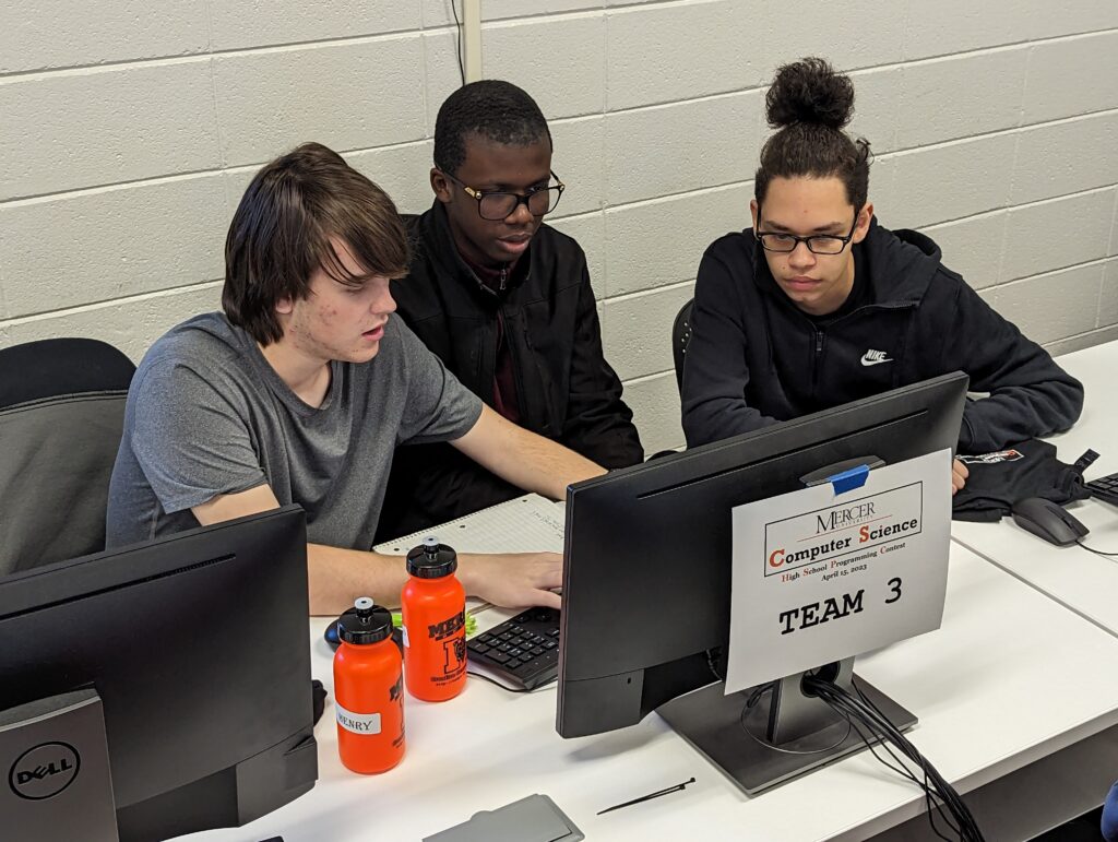 Three students are seated around a computer.