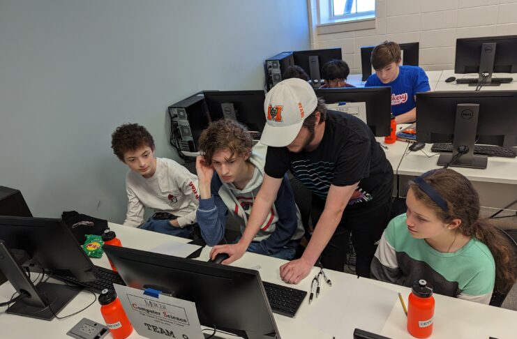 A Mercer student leans over a desk to help a team of students at their computer.