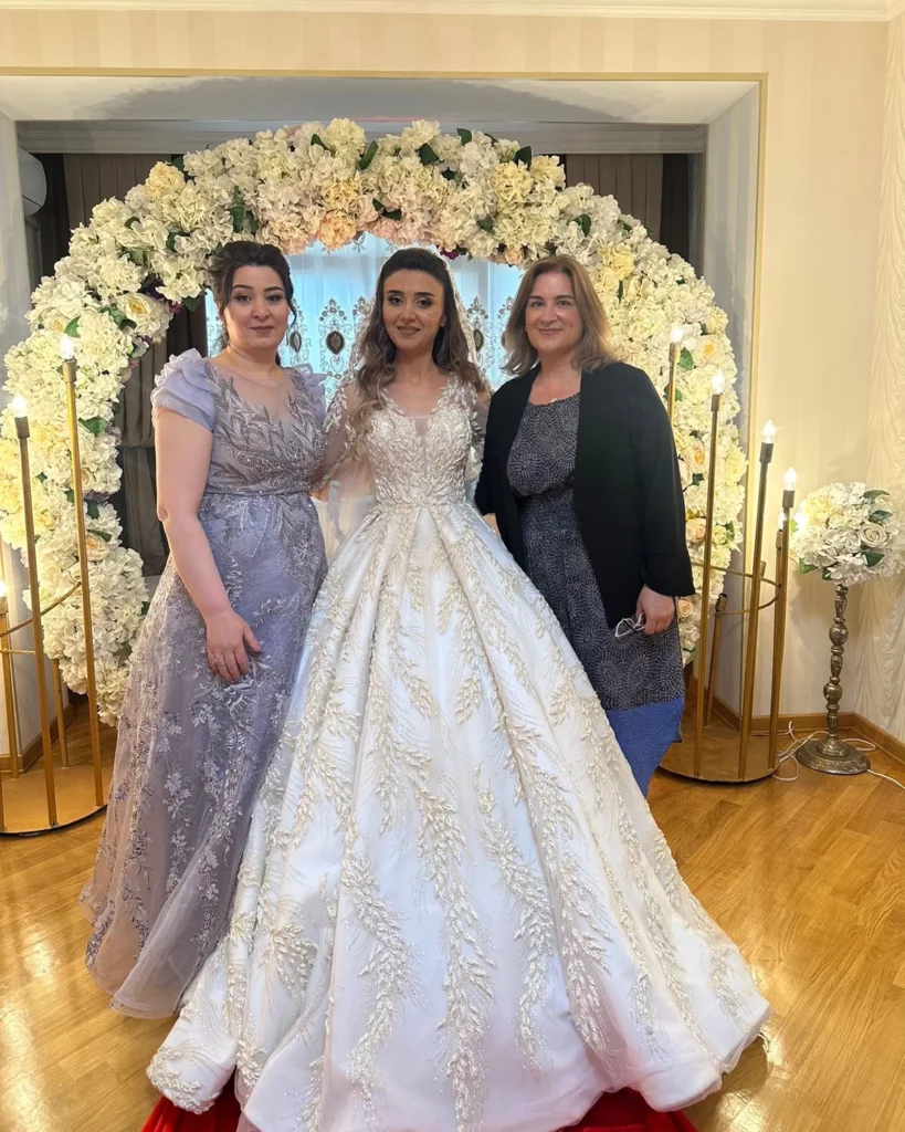 A bride in a wedding grown stands with two other women by her side and a large floral wreath behind them.