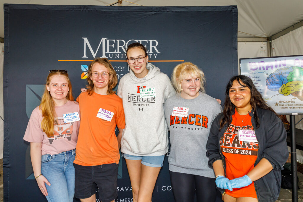 Three women stand together with arms around each other's back, in front of a Mercer backdrop