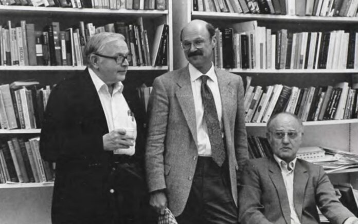 Three men stand in front of a shelf of books.