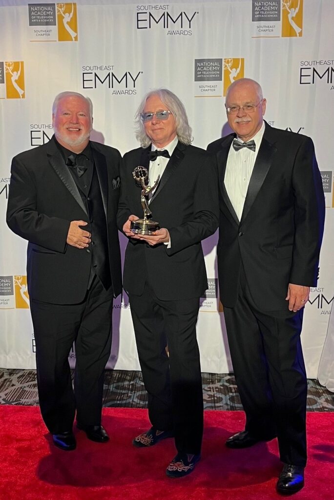 Mark Maness, Mike Mills and Larry Brumley stand in front of an Emmy backdrop, with Mills holding an Emmy statuette.