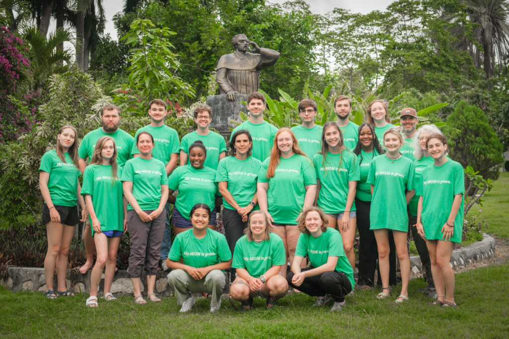 A group of students and faculty stand together, wearing matching green shirts, with a statue behind them.