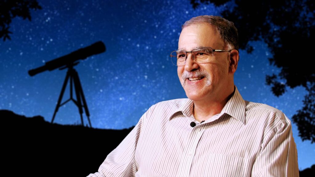 a man sits in front of a backdrop with stars and a telescope