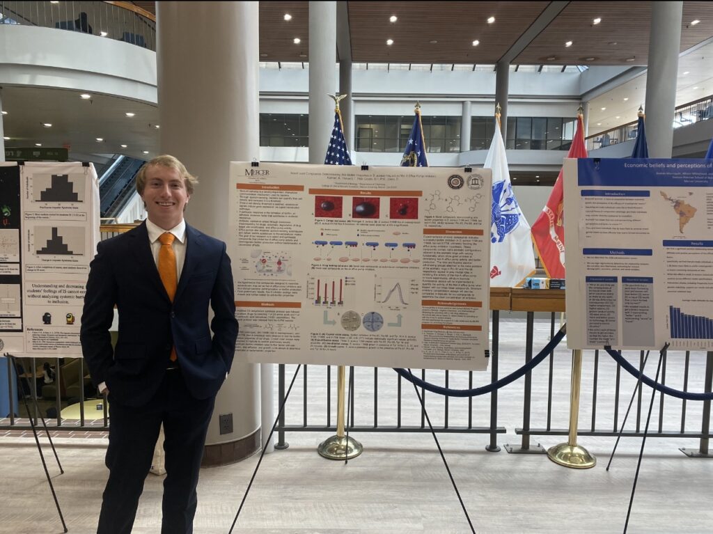 A student wears a suit and stands beside his research poster.