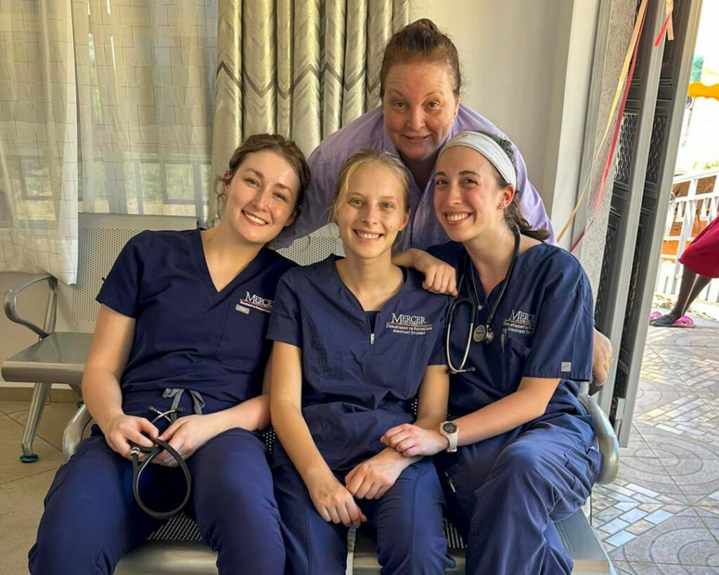 three students sit wearing blue scrubs and a woman stands behind them