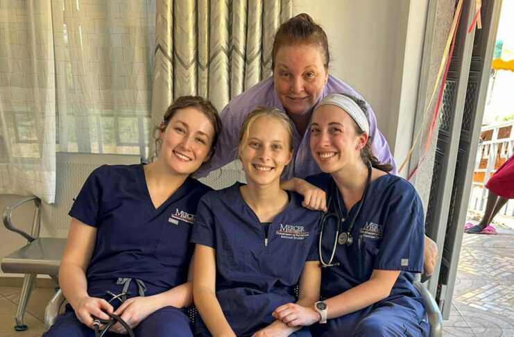 three students sit wearing blue scrubs and a woman stands behind them