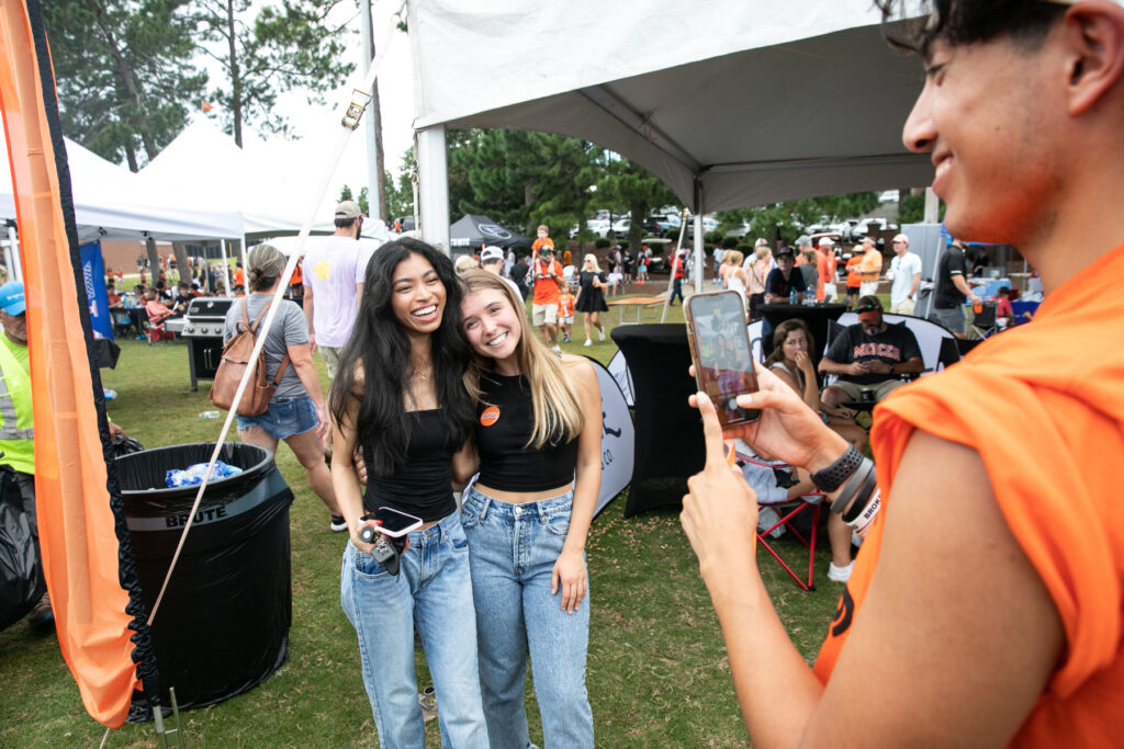 two girls in black shirts and jeans pose for a picture amongst tailgaters