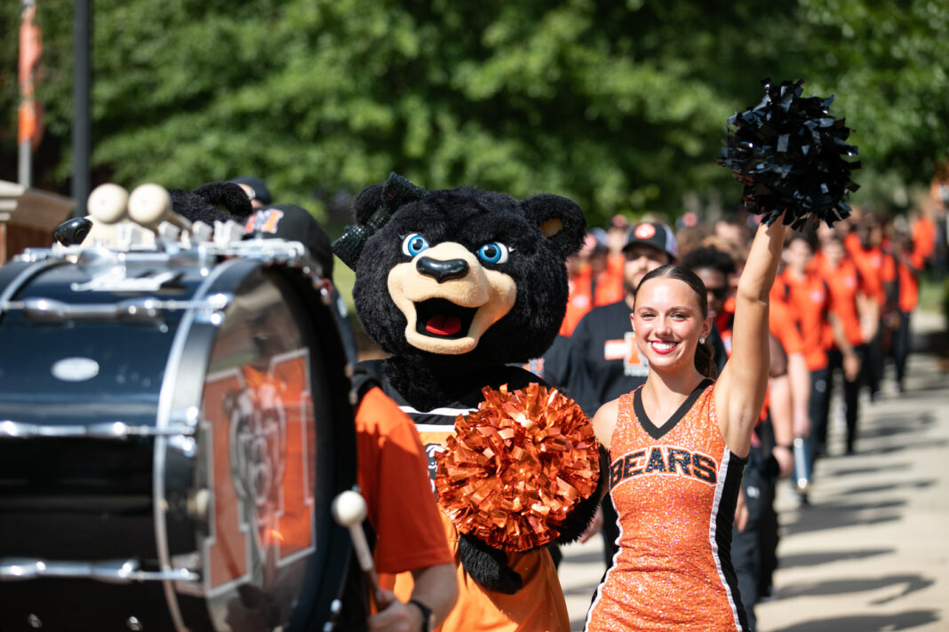 a marching band member hits a bass drum, followed by a female bear mascot and cheerleader