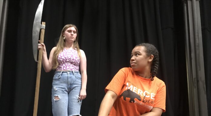Join Mercer Players for a ‘captivating and diverse journey’ down the rabbit hole A woman holds an axe over a woman kneeling at a bench during a play rehearsal.