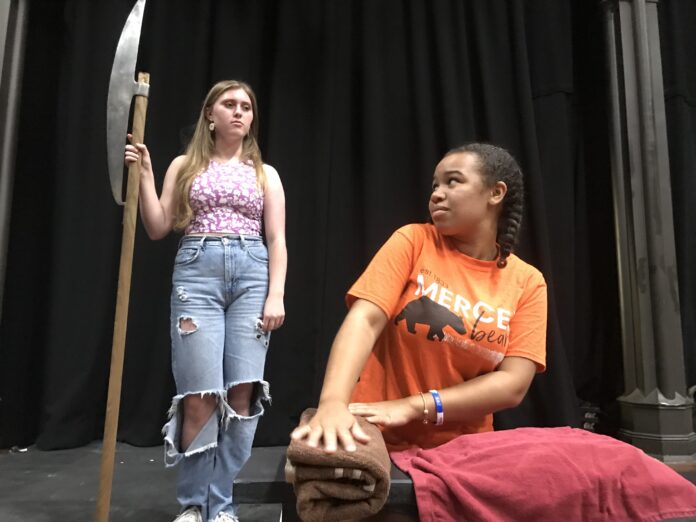 A woman holds an axe over a woman kneeling at a bench during a play rehearsal.