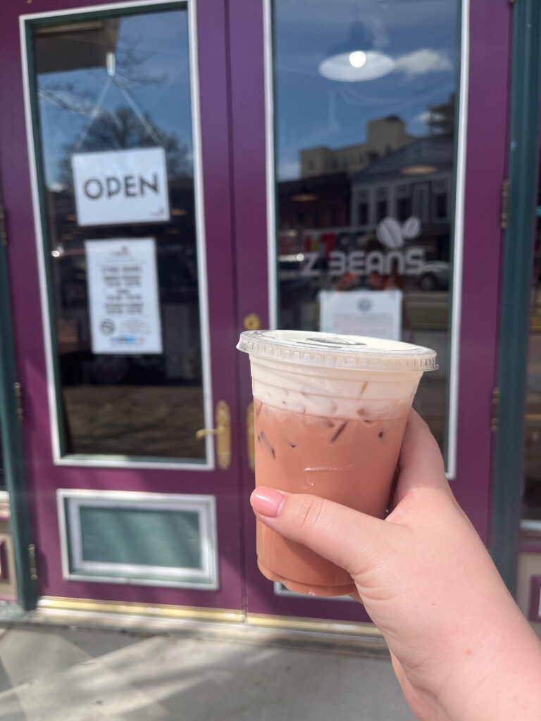 A hand is seen holding a cold drink in front of the doors to Z Beans.