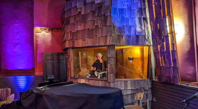 Professor records album examining illness and grief at Capricorn Sound Studios A man sings inside a studio both, wearing headphones and holding his guitar.