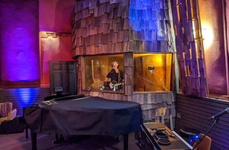 A man sings inside a studio both, wearing headphones and holding his guitar.