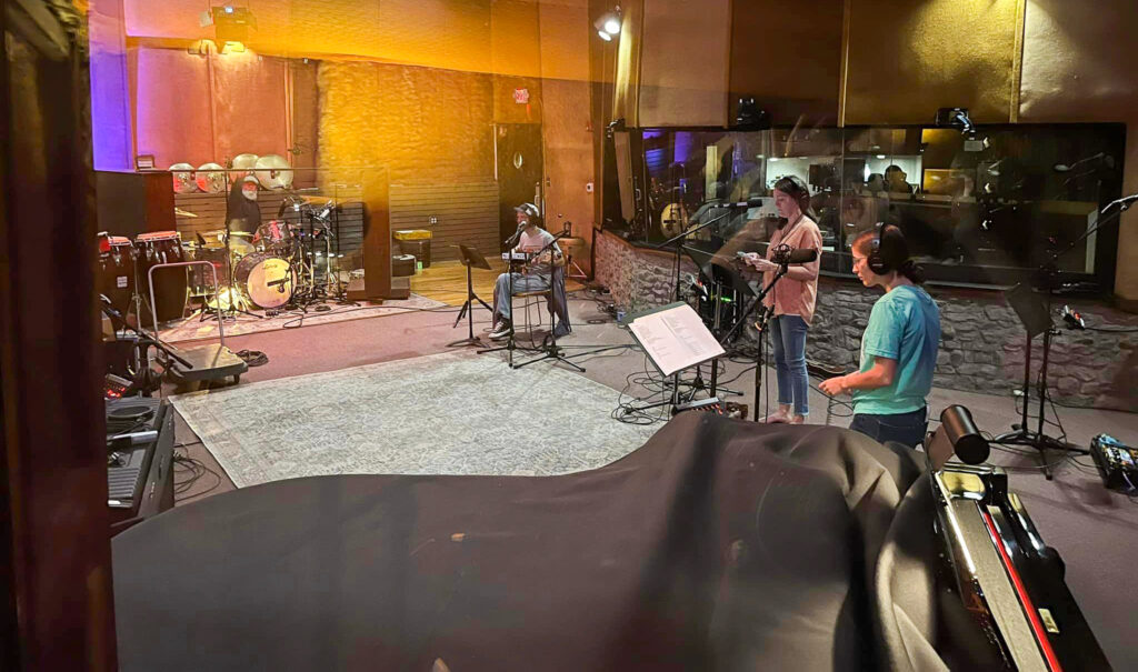 Musicians on drums and guitar and two vocalists perform inside a studio.