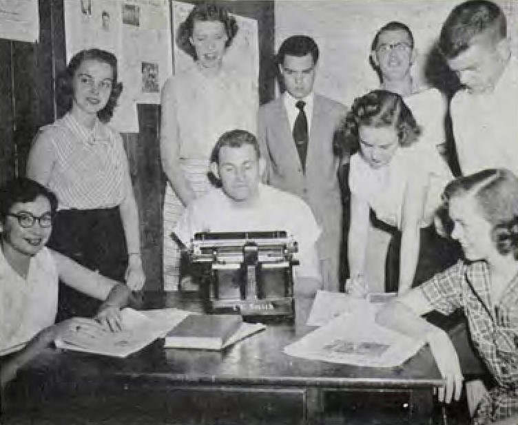 Several people gather around a man typing at a typwriter.