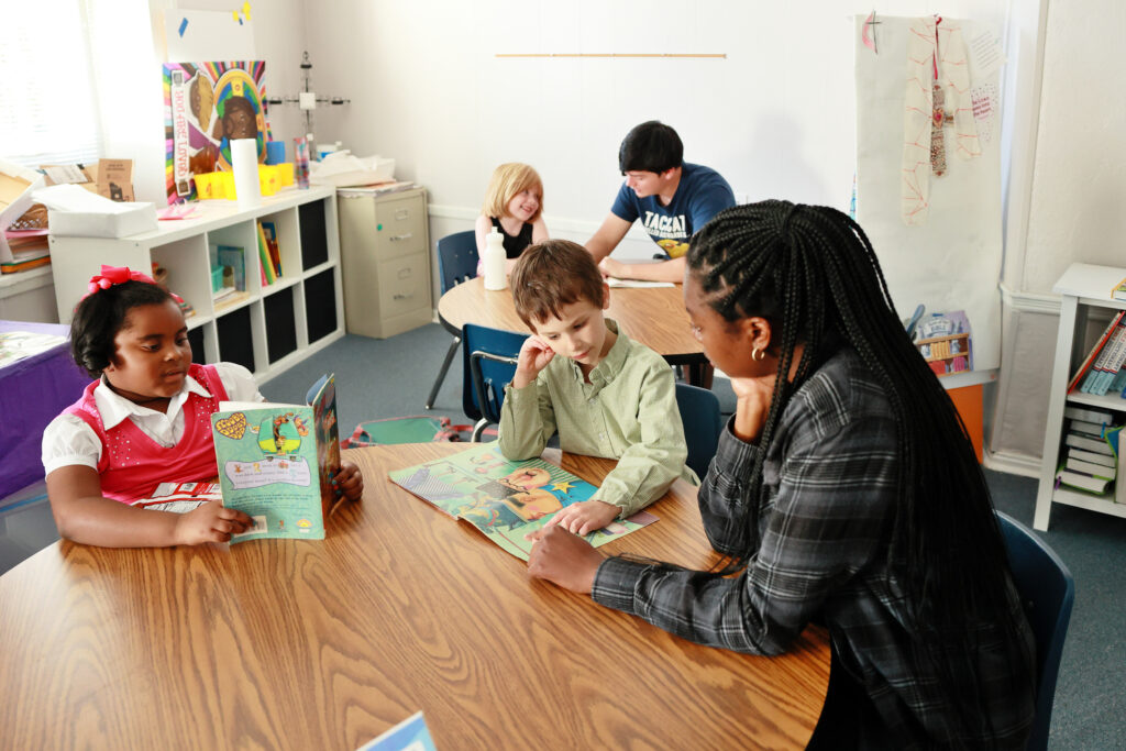 Two college students sit at tables with two children to practice reading skills.