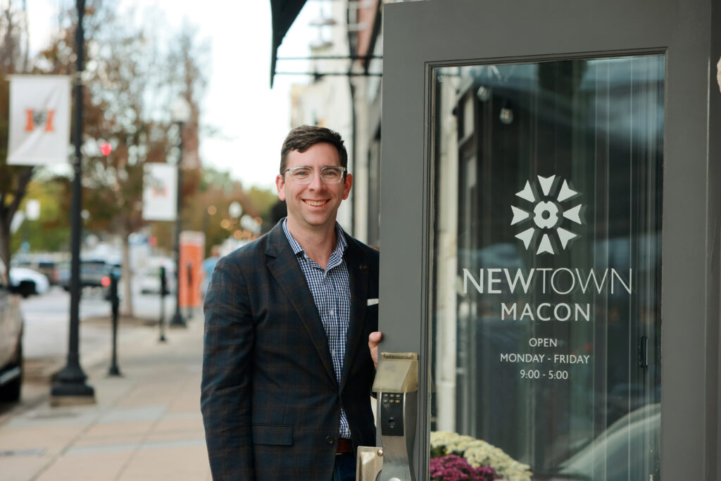 A man in a suit holds a door open that says "NewTown Macon," with a view of downtown Macon behind him.