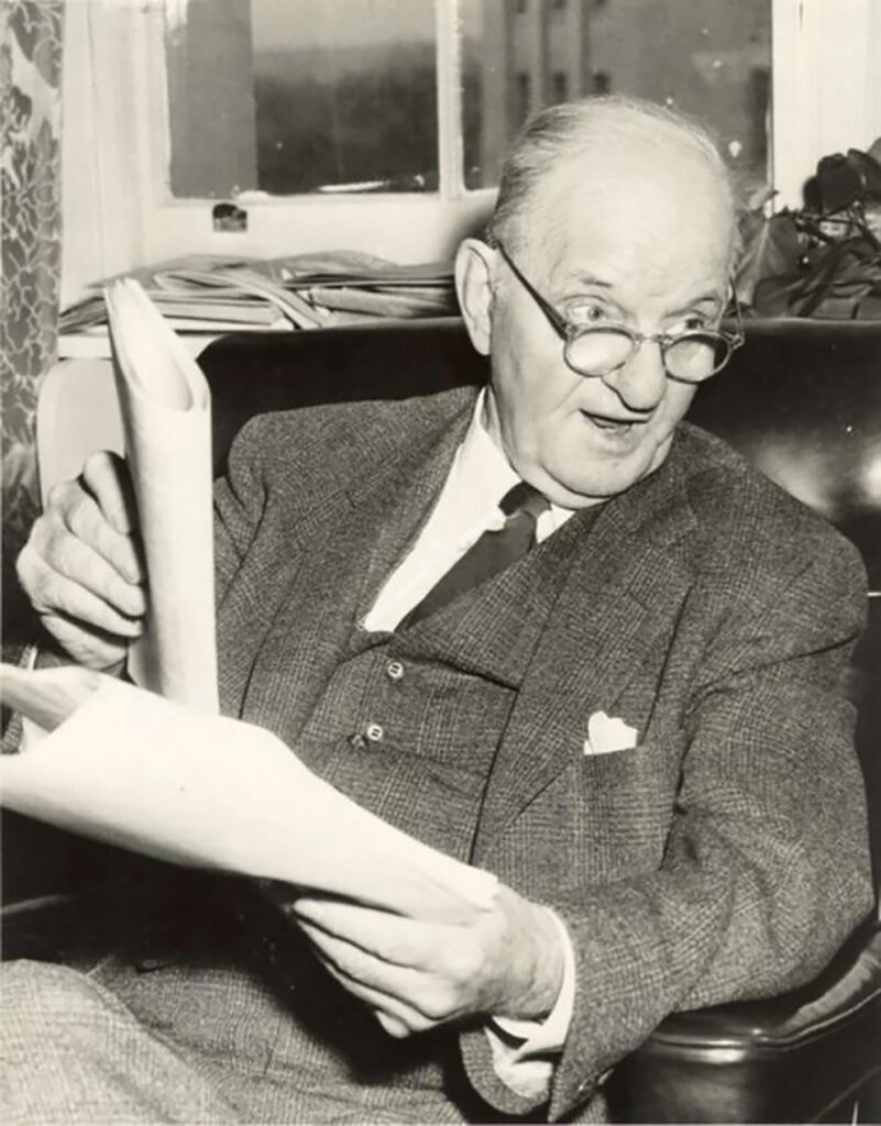a black and white photo of a man in a suit sitting in a leather chair and holding papers