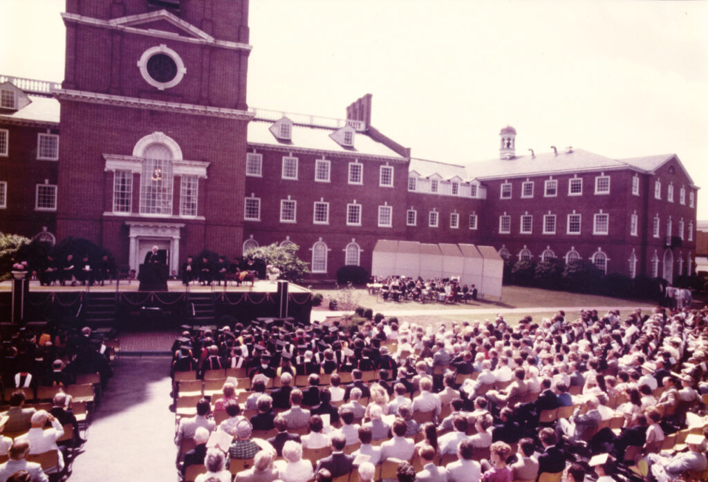 a man speaks on stage in front of the a large brick building. a crowd looks on
