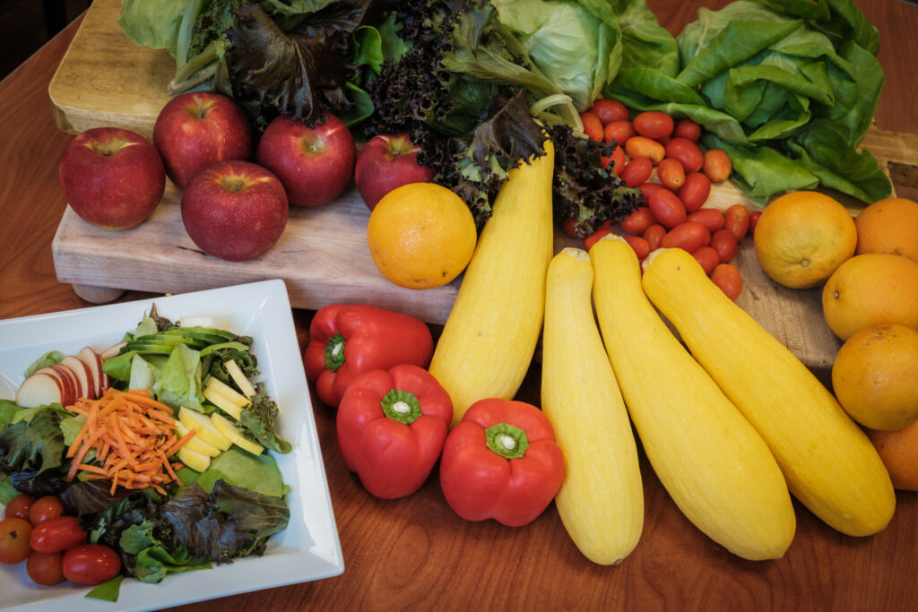 an array of fresh produce including lettuce, apples, red bell peppers, yellow squash, oranges and a side salad