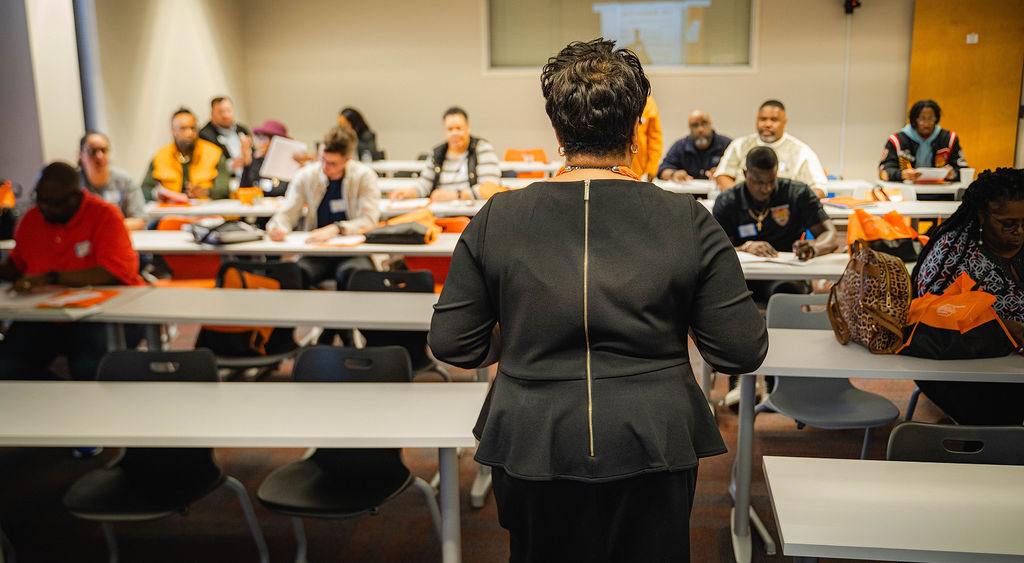 A woman teaches at the front of the room while her students sit at long tables in front of her.