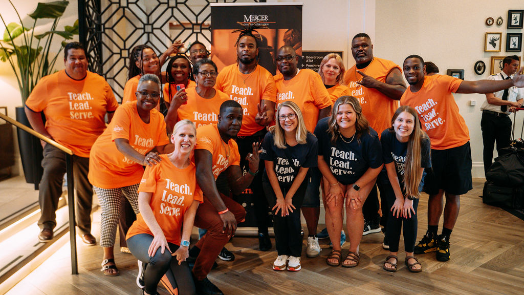A group of people wearing orange and black shirts that say "Teach. Lead. Serve" post for a photo.