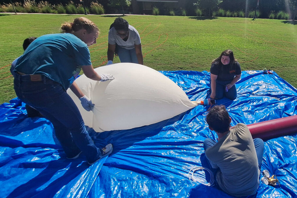 students on a blue tarp blow up a partially deflated white balloon