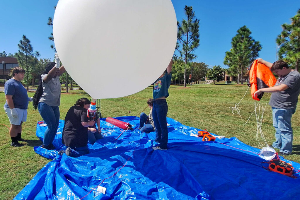 students on a blue tarp blow up a white balloon