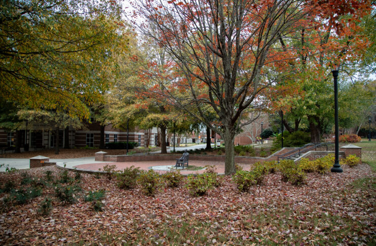Park scene with autumn leaves, a circular seating area with benches, brick pathway, trees with orange leaves, and a lamppost.