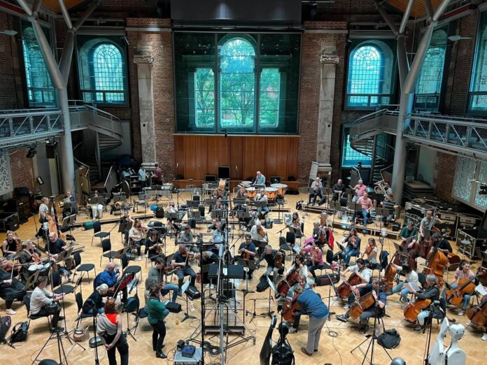 Orchestra musicians can be seen at their seats with their instruments, in a large open venue with large windows behind them.