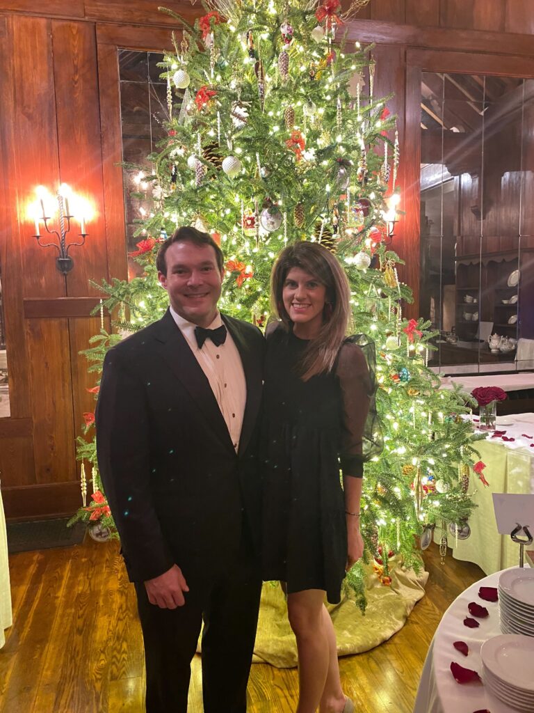 A man and woman stand in front of a Christmas tree, dressed in nice outfits. 