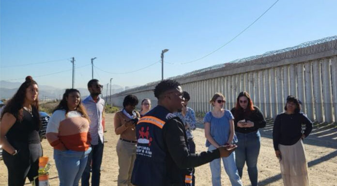 French and Spanish students participate in service-learning at U.S. border a group of people listen to a man talking. a wall with barbed wire is in the background