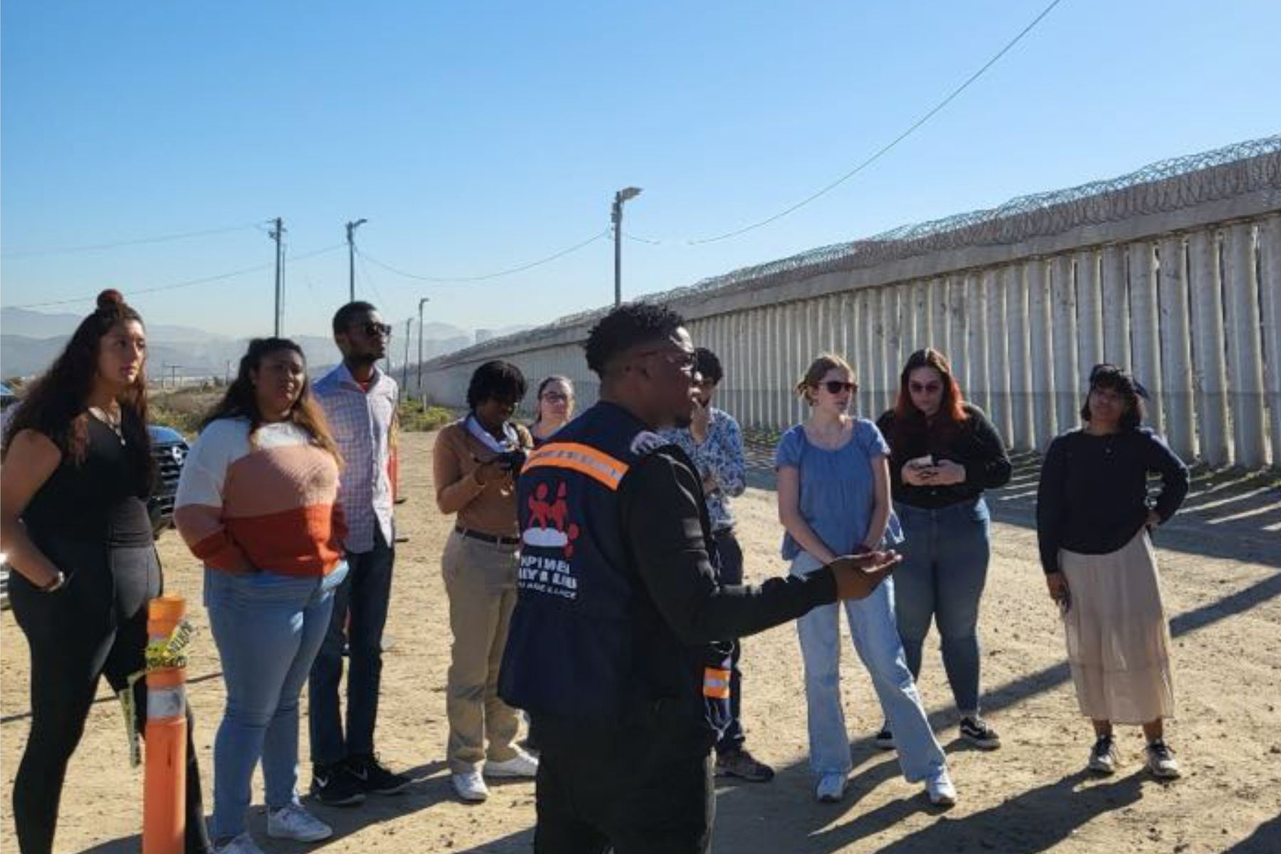 a group of people listen to a man talking. a wall with barbed wire is in the background