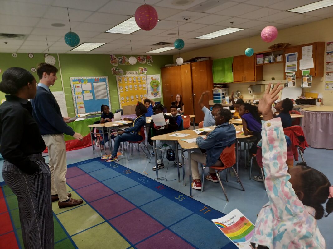 Two adults stand in the front of a classroom, with third-graders at tables looking at them and several raising their hands.