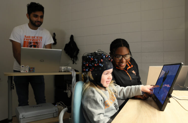 Parth Patel stands at a computer, while Lynzi Holland sits at a table with a child wearing a cap with the fNIRS technology.