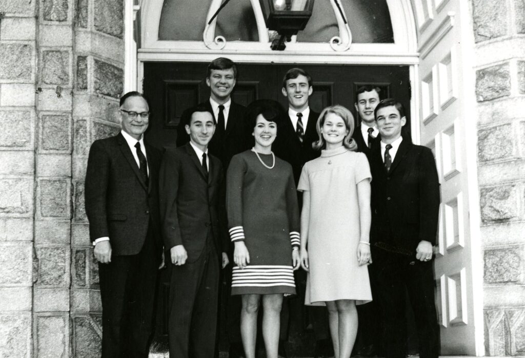 A black and white photo of eight people standing on steps in front of the door of a brick building.