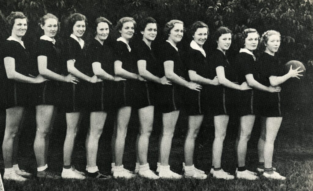 A black and white photo of several women in black shirts and shorts stand in a line with their hands around each other's waists, with the last woman on the right holding a basketball.