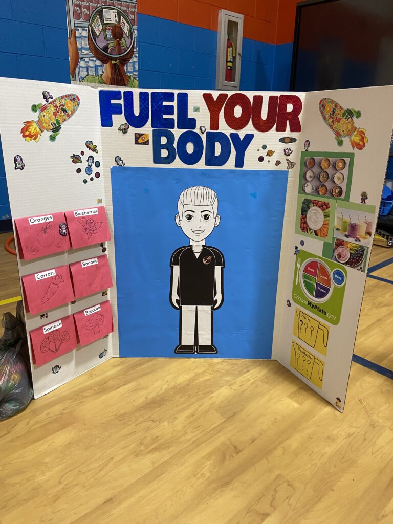 A posterboard is titled "Fuel Your Body" and shows a cartoon of a boy in the middle and fruits and vegetables on the sides.